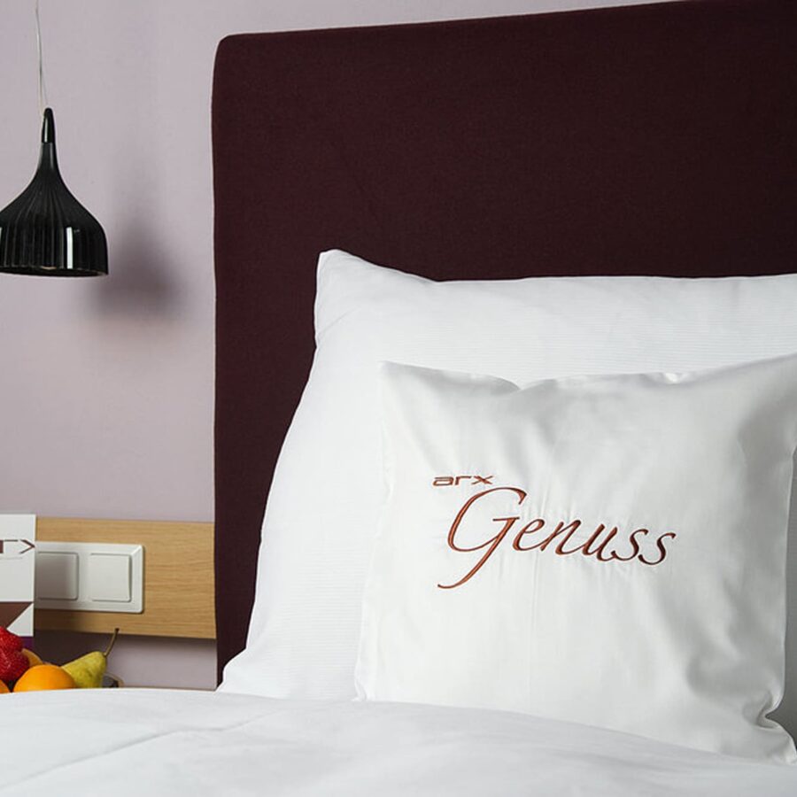 Hotelbett mit weißem Kissen bestickt mit arx Genuss, dunklem Kopfteil, schwarzer Hängelampe und Obstschale auf, Referenzen