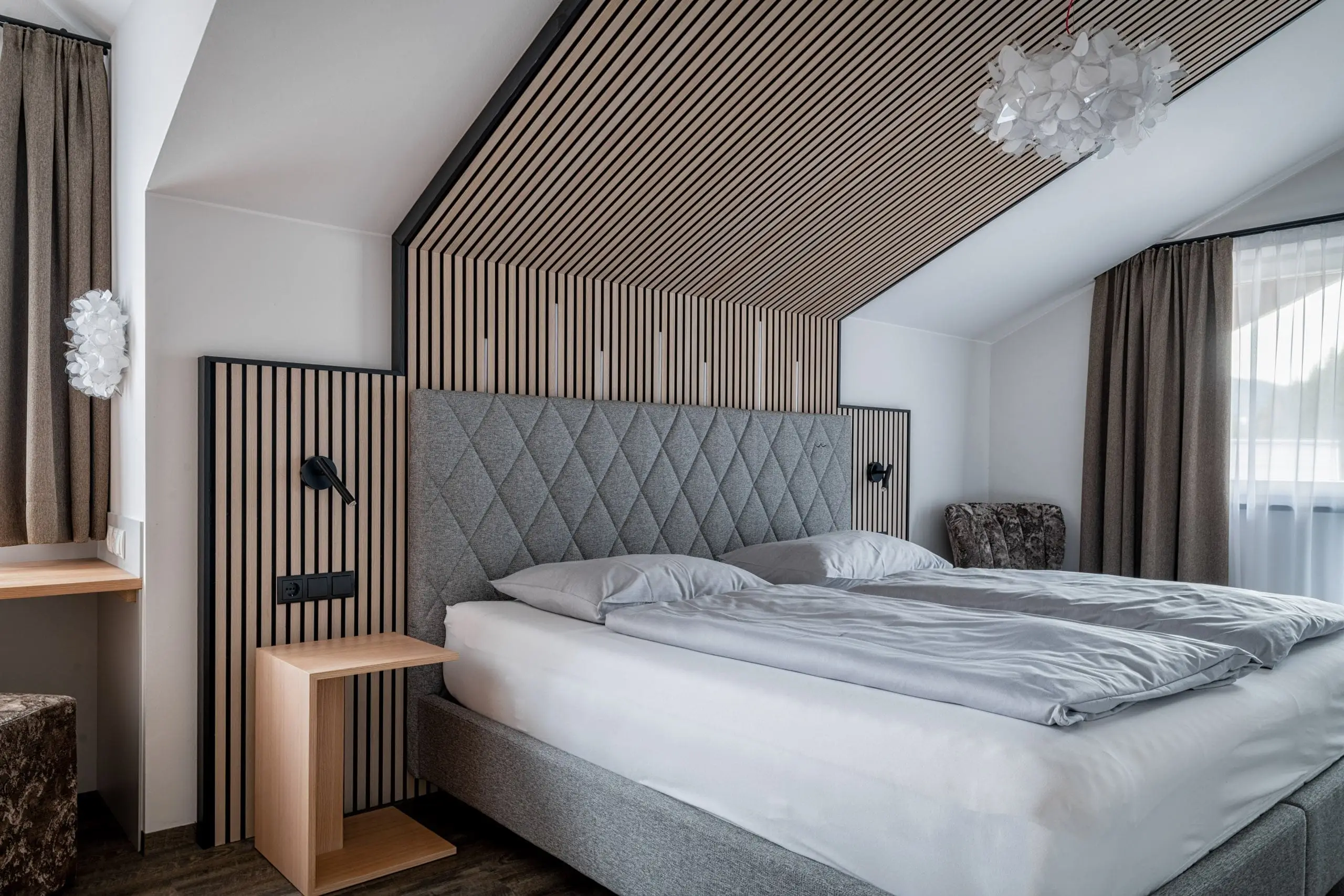 Modernes Schlafzimmer mit maßgefertigtem Kastenverbau aus Holzlamellen, grauem Polsterbett, Leselampen und, Alpenblick