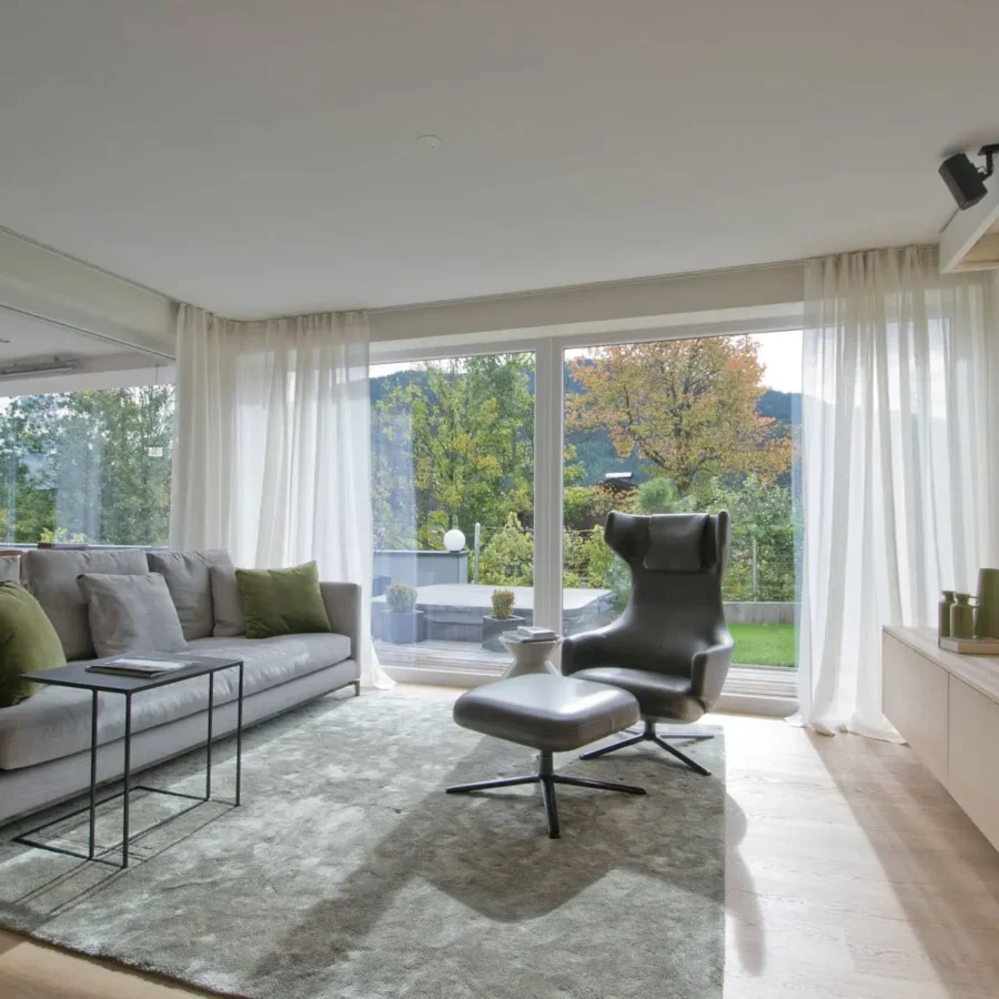 Moderner Wohnraum mit grauem Sofa, grünem Kissen, schwarzem Ledersessel mit Hocker, großem Fenster und, Referenz