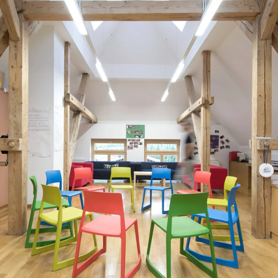 Moderner Schulraum mit bunten Vitra Tip Ton Stühlen im Kreis, sichtbare Holzbalken und helle Innenarchitektur, Referenz