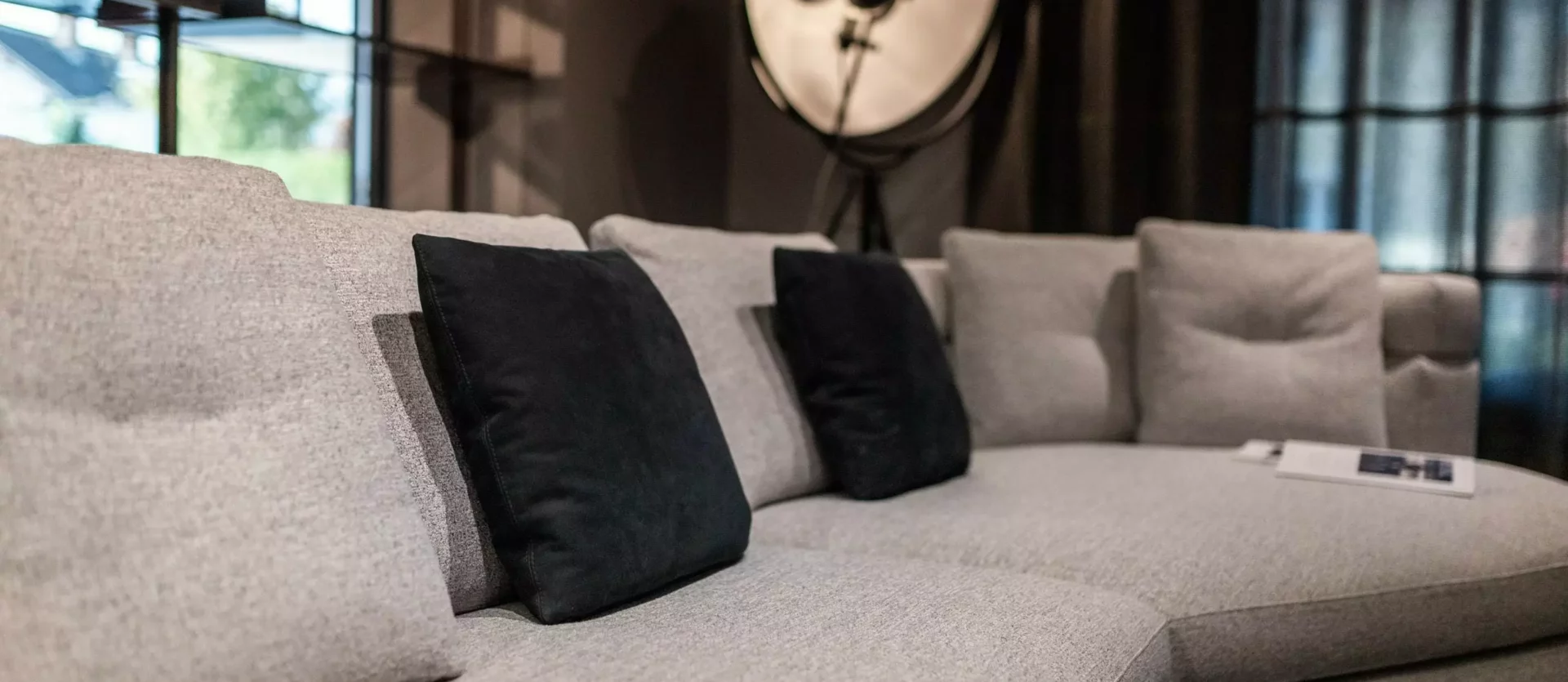 Großes graues Polstermöbel mit schwarzen Kissen vor Fenster und Stehlampe im modernen Möbelstudio mit, Poliform
