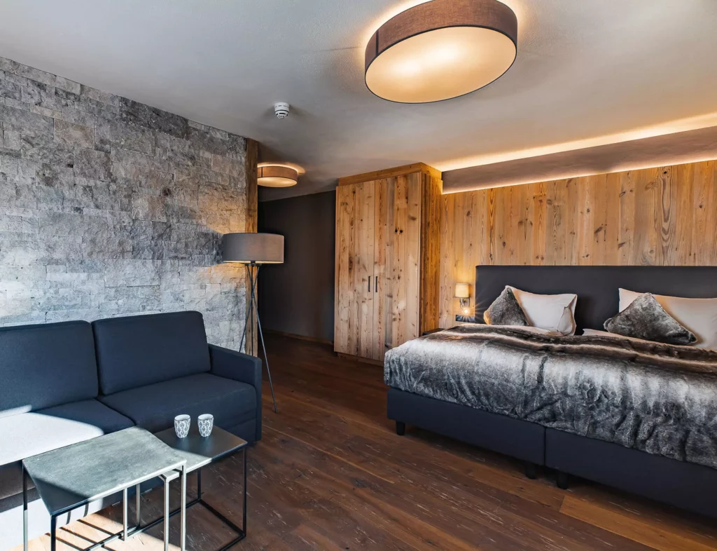 Modern eingerichtetes Hotelzimmer mit Altholz-Wandverkleidung, grauem Boxspringbett, Sofa, Stehlampe und, Allmonte