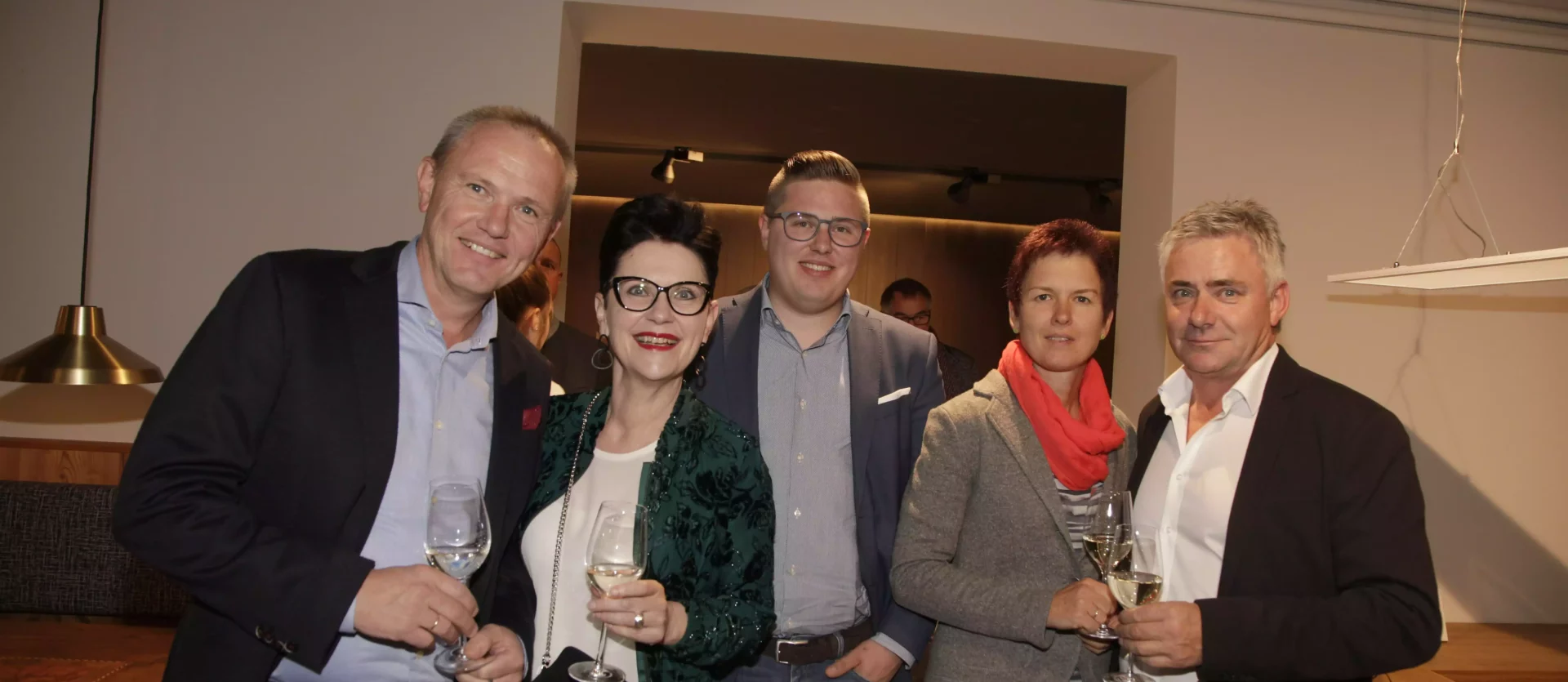 Fünf Personen in eleganter Kleidung mit Weingläsern in einem modernen Showroom der Tischlerei und Innenarchitektur, Fest