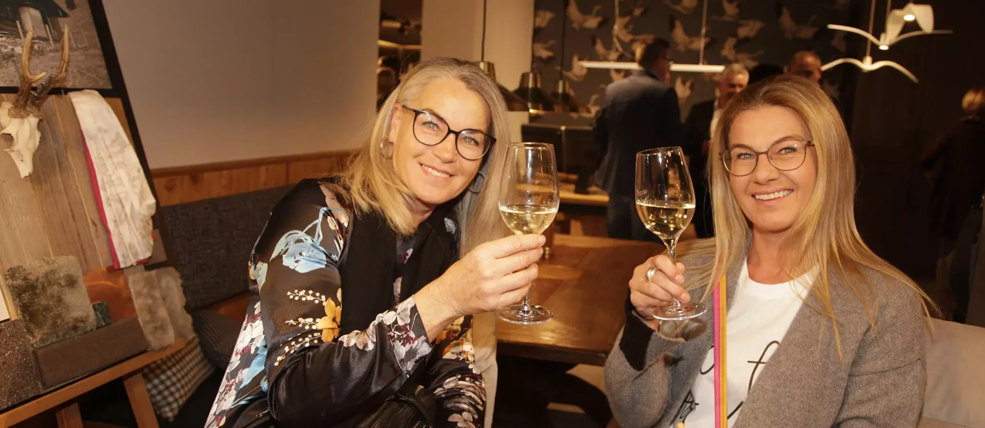 Zwei Frauen mit Brillen stoßen mit Weißweingläsern in einem gemütlichen Möbelstudio mit Holztisch und, Fest