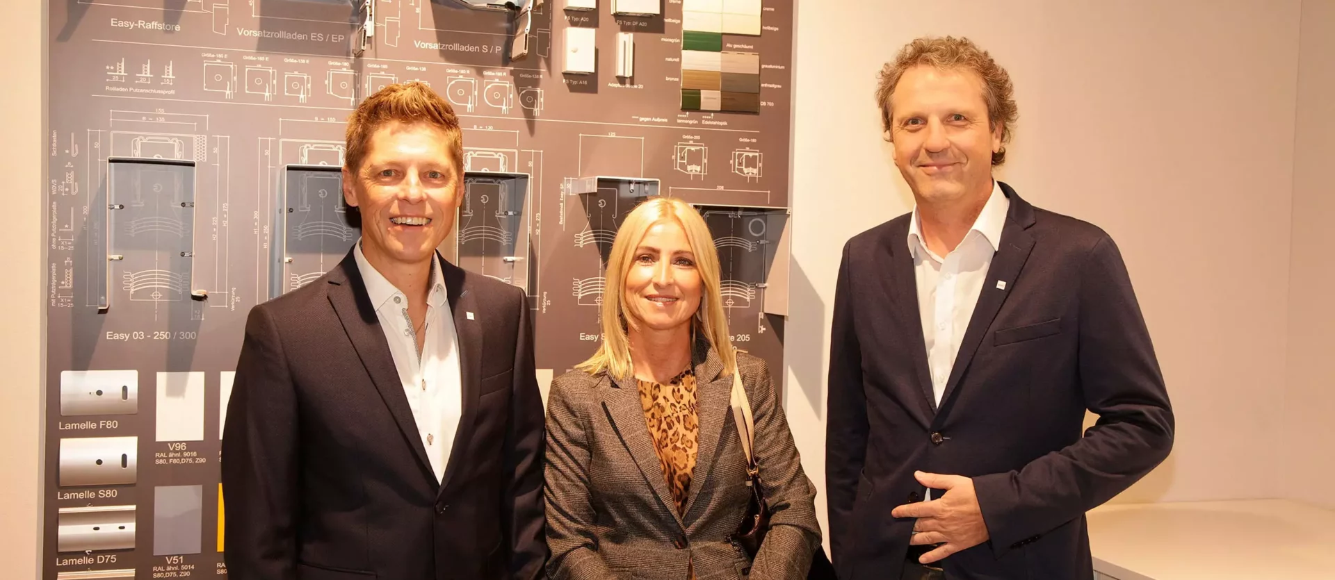 Drei Personen in Businesskleidung stehen vor einer Wand mit technischen Zeichnungen und Mustern von Rollladenlamellen, Fest