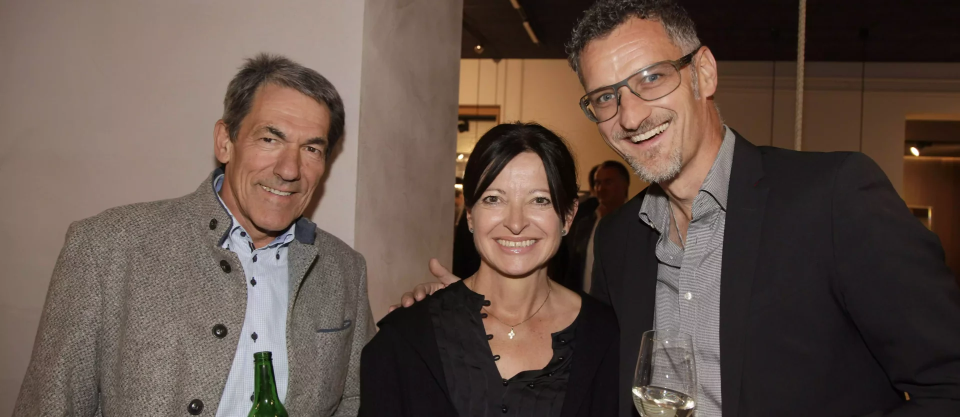 Drei Personen in einem Innenraum bei einem Event, zwei Männer und eine Frau, halten Getränke, freundliche Atmosphäre, Fest