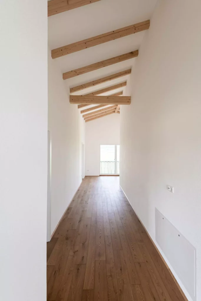 Langer heller Flur mit sichtbaren Holzbalken an der Decke und braunem Holzboden, weiße Wände und Fenster am Ende, Rimadesio
