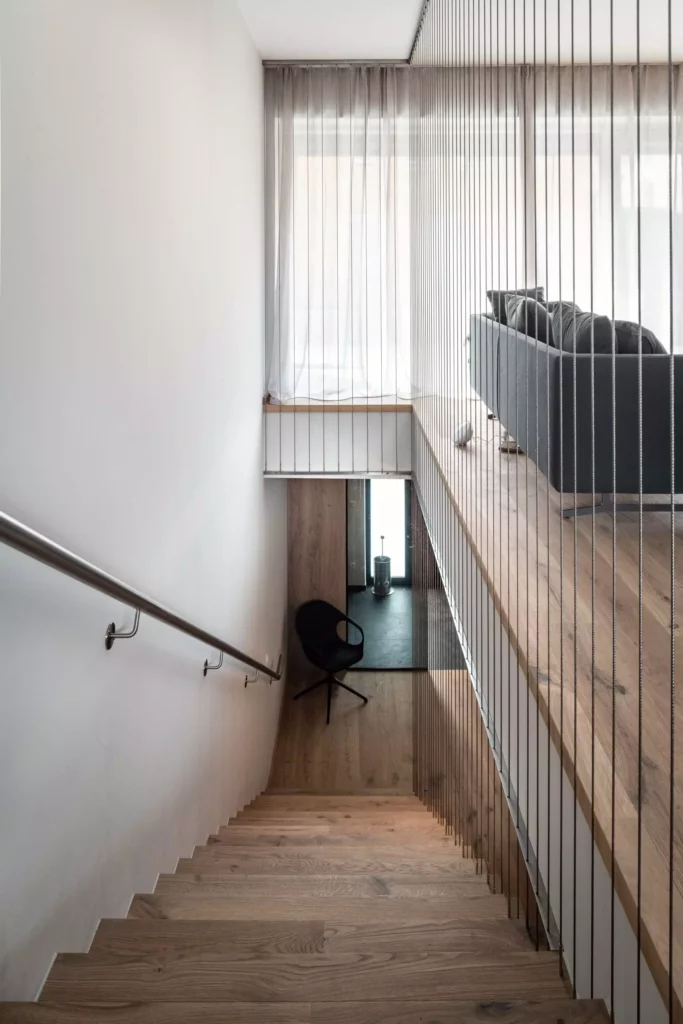 Moderner Treppenaufgang mit Holzstufen, Edelstahlhandlauf, schwarzem Stuhl unten und offener Galerie mit grauem, Möbelstudio