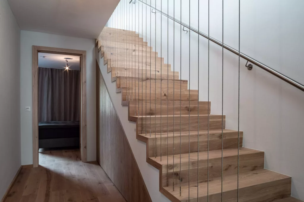 Moderne Holztreppe mit Edelstahlseilgeländer und Handlauf neben Flur mit Holzboden und offener Tür zu, Möbelstudio