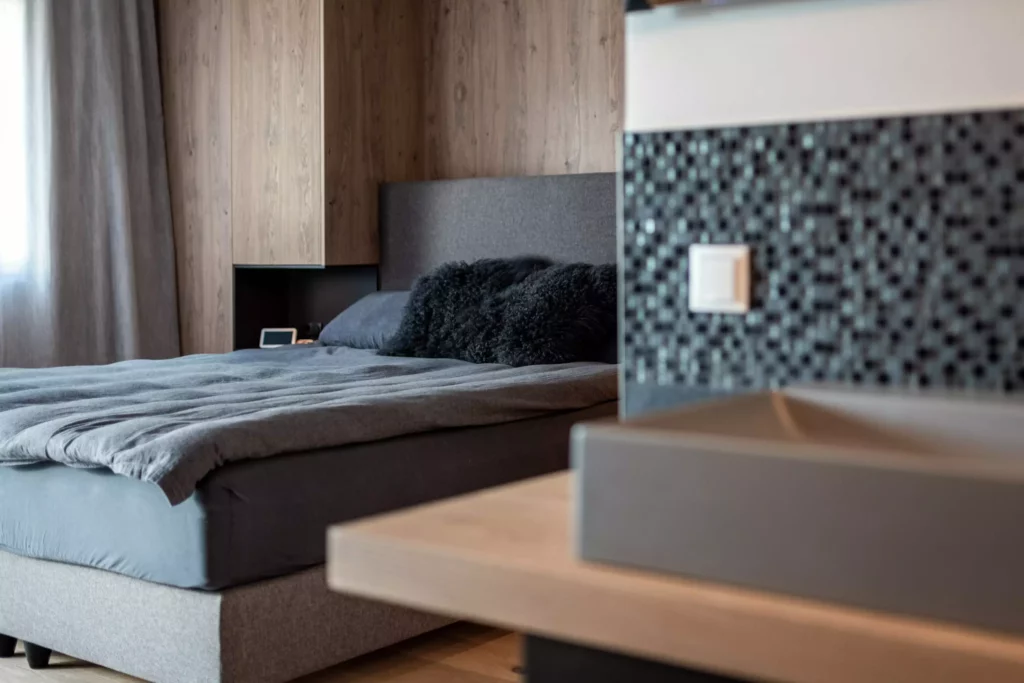 Modernes Schlafzimmer mit grauem Polsterbett, schwarzen Kissen, Holzverkleidung und Mosaikfliesen im Vordergrund, Möbelstudio