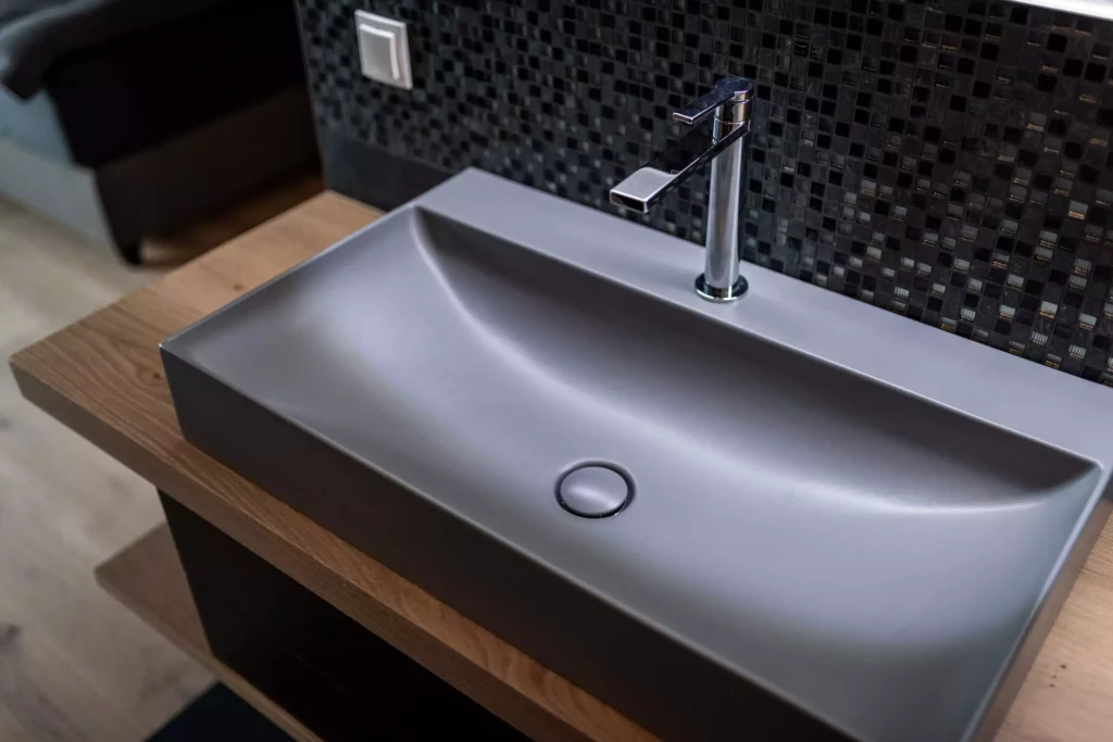 modernes graues Waschbecken mit Chromarmatur auf Holzplatte vor schwarzer Mosaikwand in maßgefertigtem, Möbelstudio