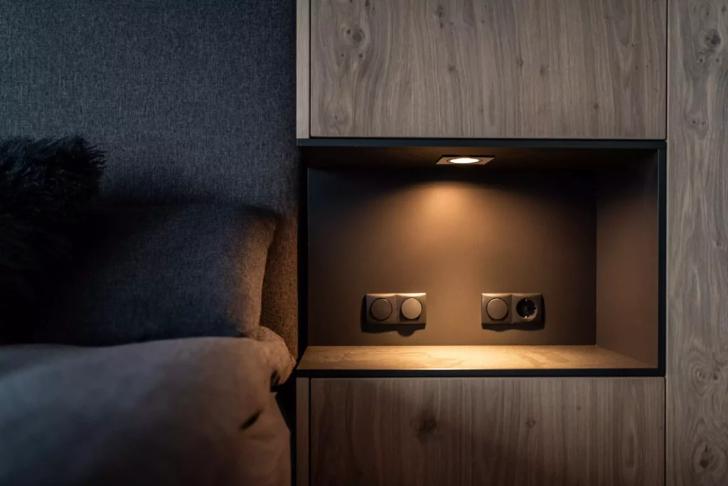 Nische mit indirekter Beleuchtung, Steckdosen und Schaltern neben einem dunkelgrauen Bett mit Kissen, Möbelstudio