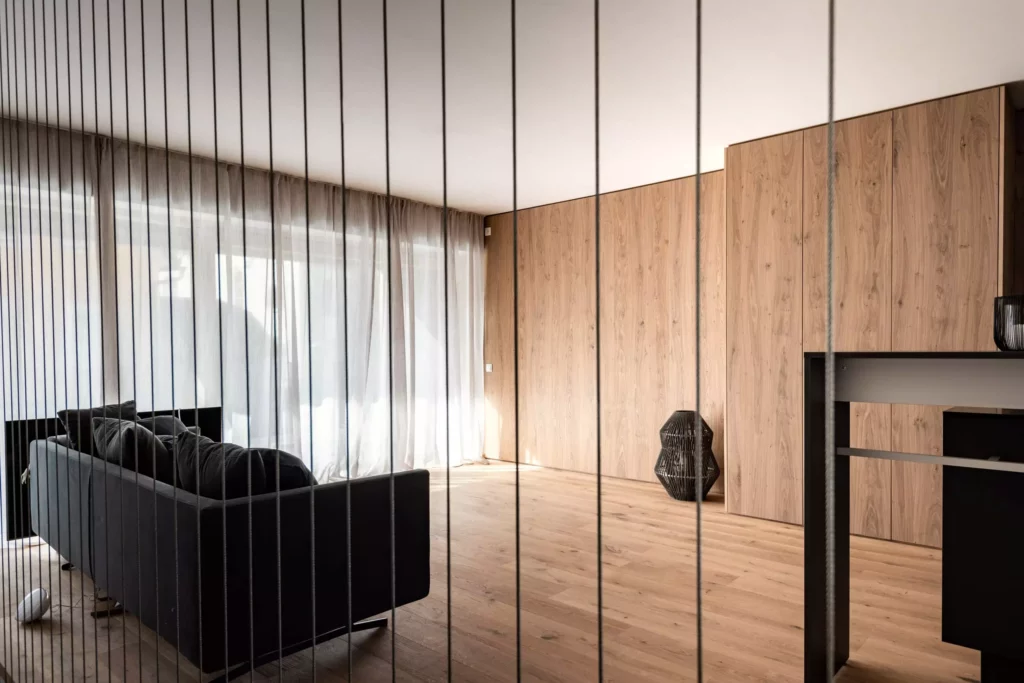 Moderner Wohnraum mit maßgefertigten Holzpaneelen, schwarzem Sofa, großem Fenster mit transparenten Vorhängen, Möbelstudio