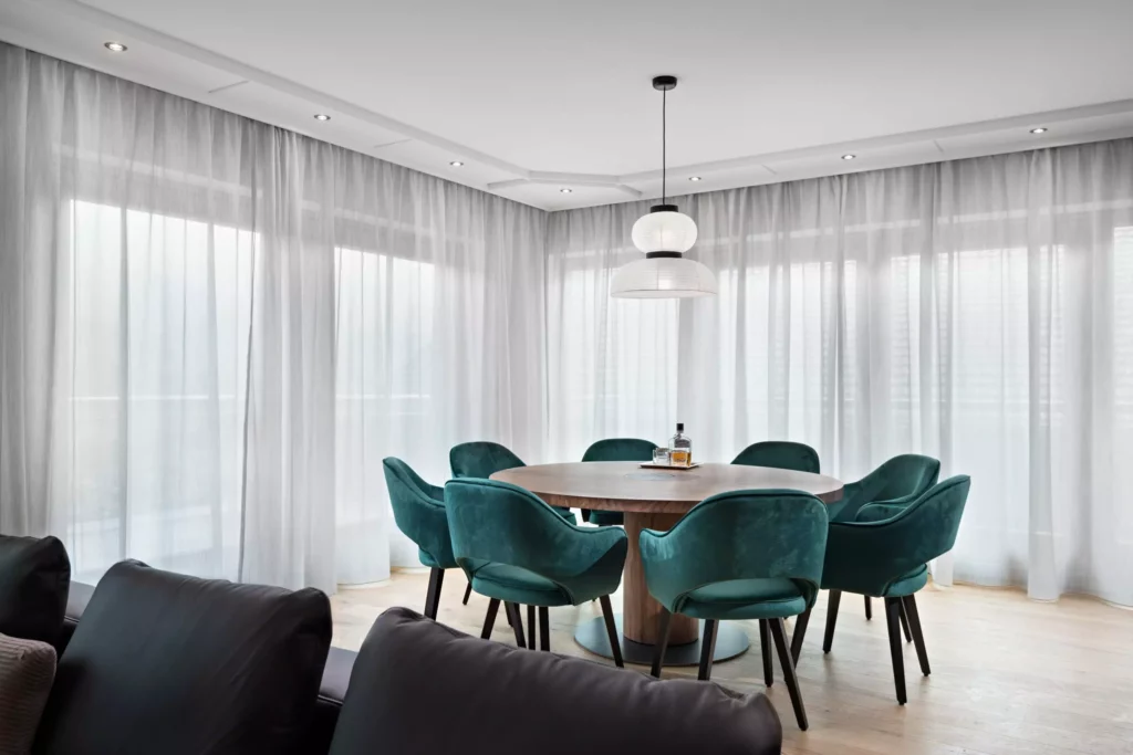 Moderner Essbereich mit rundem Holztisch, sechs grünen Samtstühlen und großer Fensterfront mit weißen Vorhängen in, Riva1920
