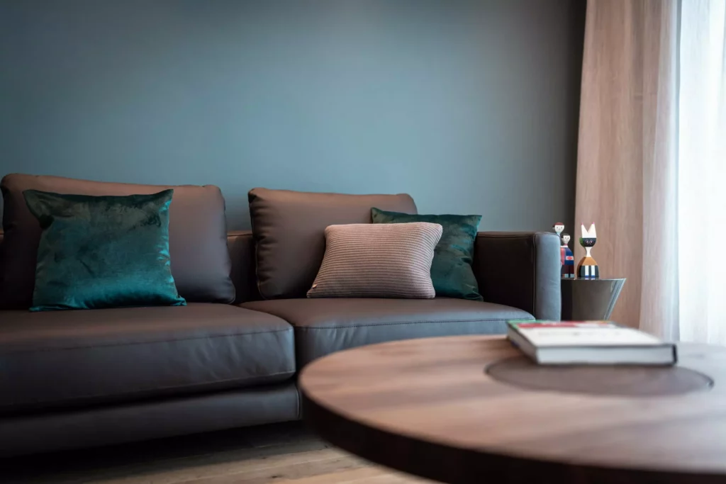 Modernes Wohnzimmer mit braunem Ledersofa, grünen und grauen Kissen, rundem Holztisch und dekorativen Figuren, Riva1920