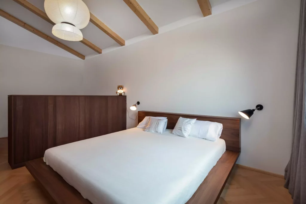 Schlafzimmer mit maßgefertigtem Holzbett und Raumteiler aus dunklem Holz, Wandlampen und Holzdeckenbalken in, Riva1920