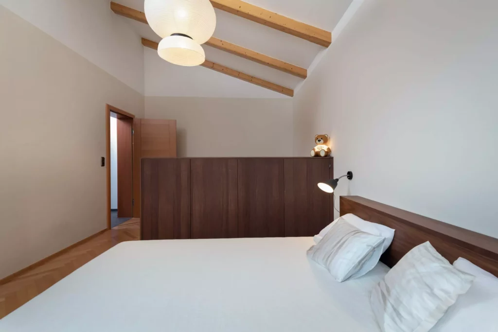 Schlafzimmer mit maßgefertigtem Holzbett und Raumteiler, weiße Wände, Holzdecke mit sichtbaren Balken, Teddy-Lampe, Riva1920