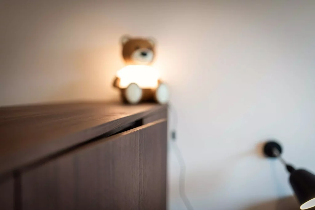 Nahaufnahme einer dunklen Holzkommode mit beleuchtetem Teddybär und moderner Wandlampe im Hintergrund, Riva1920