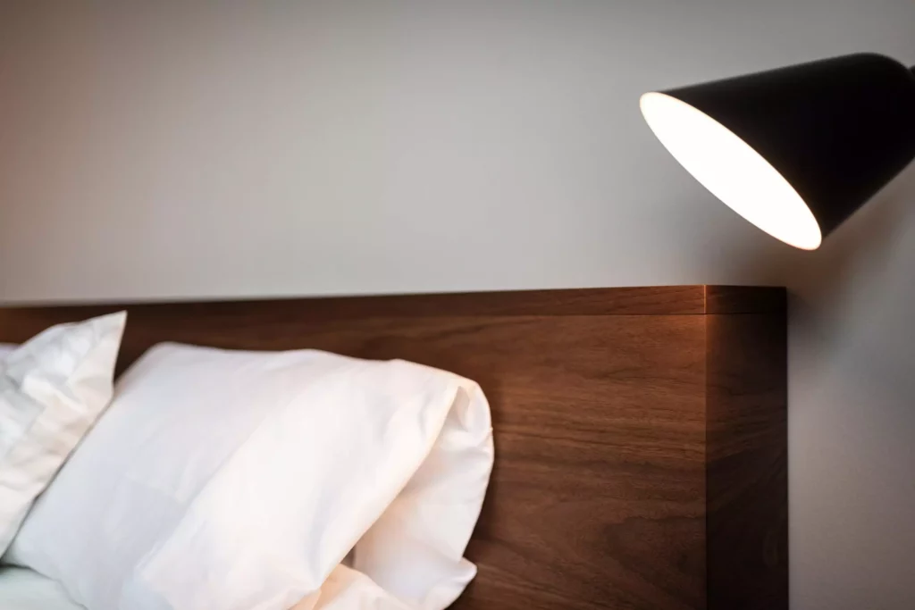 Nahaufnahme eines Betthaupts aus dunklem Holz mit weißen Kissen und einer schwarzen Wandlampe in modernem Innenraum, Riva1920