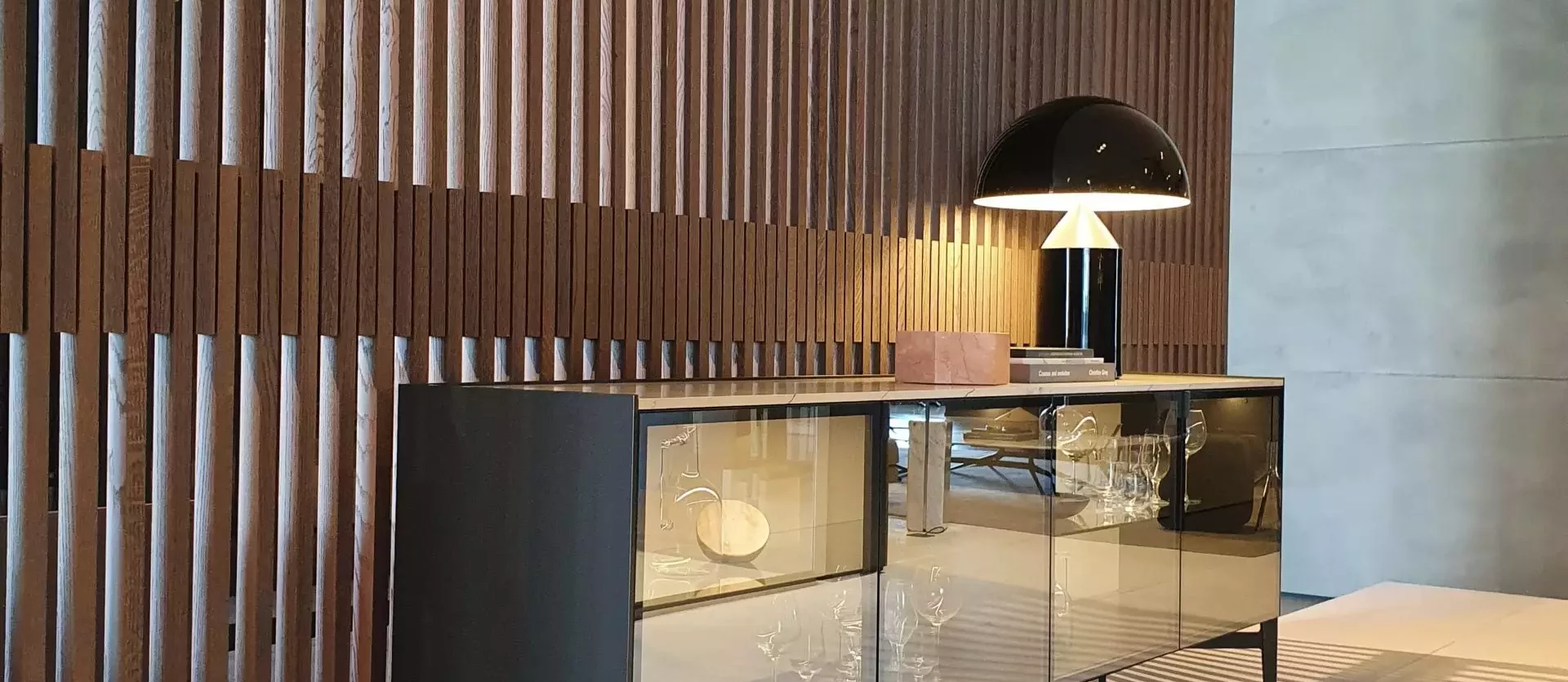 Modernes Sideboard mit spiegelnden Fronten, schwarzer Designlampe und dekorativen Gläsern vor vertäfelter Holzwand, Poliform