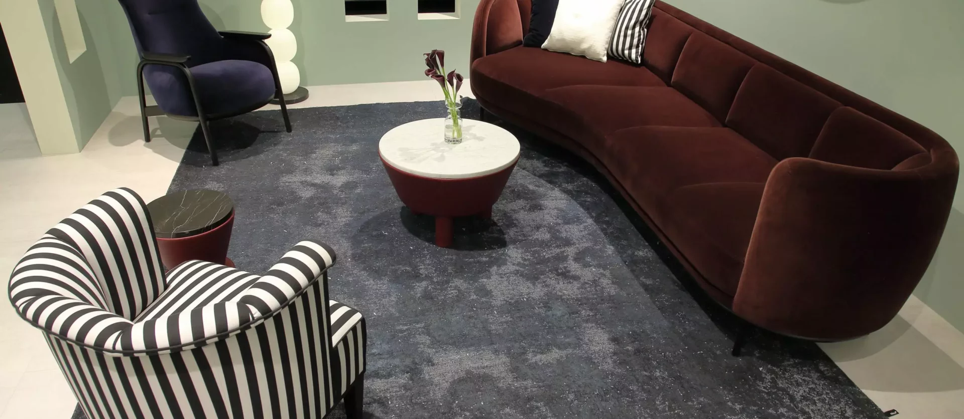 Wohnzimmer mit bordeauxrotem Wittmann Sofa, gestreiftem Sessel, dunkelblauem Sessel, runden Tischen und modernem Teppich im