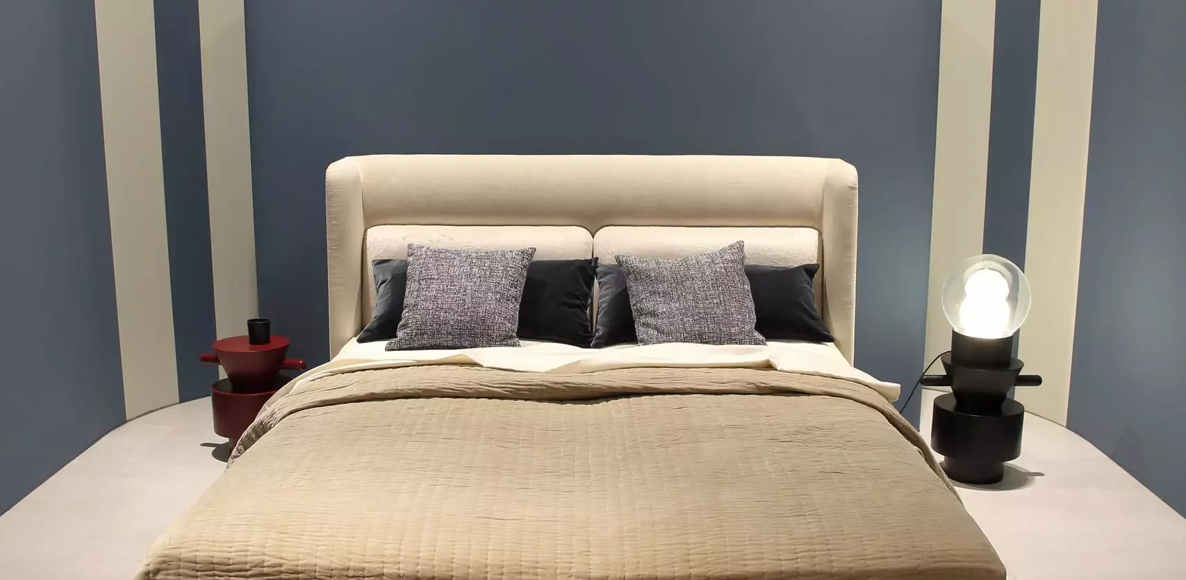 Boxspringbett der Marke Wittmann mit gepolstertem Kopfteil, brauner Bank am Fußende, zwei Nachttischen und gestreifter Wand