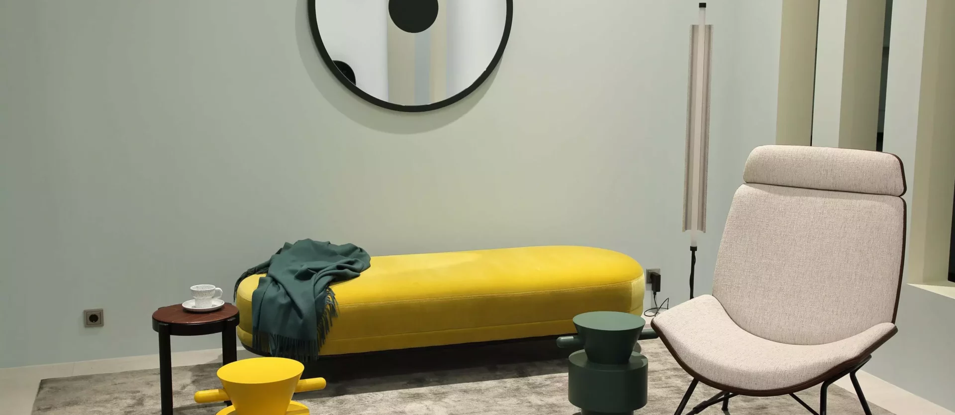Moderner Wohnraum mit gelber Bank, beigem Designersessel, zwei bunten Hockern, rundem Spiegel und Stehleuchte, Wittmann