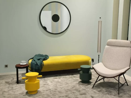 Moderner Wohnraum mit gelber Bank, beigem Designersessel, zwei bunten Hockern, rundem Spiegel und Stehleuchte, Wittmann