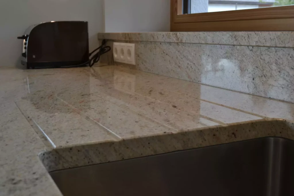 Küchenarbeitsplatte aus poliertem Naturstein mit integriertem Spülbecken und braunem Toaster vor Fensterrahmen, Referenzen