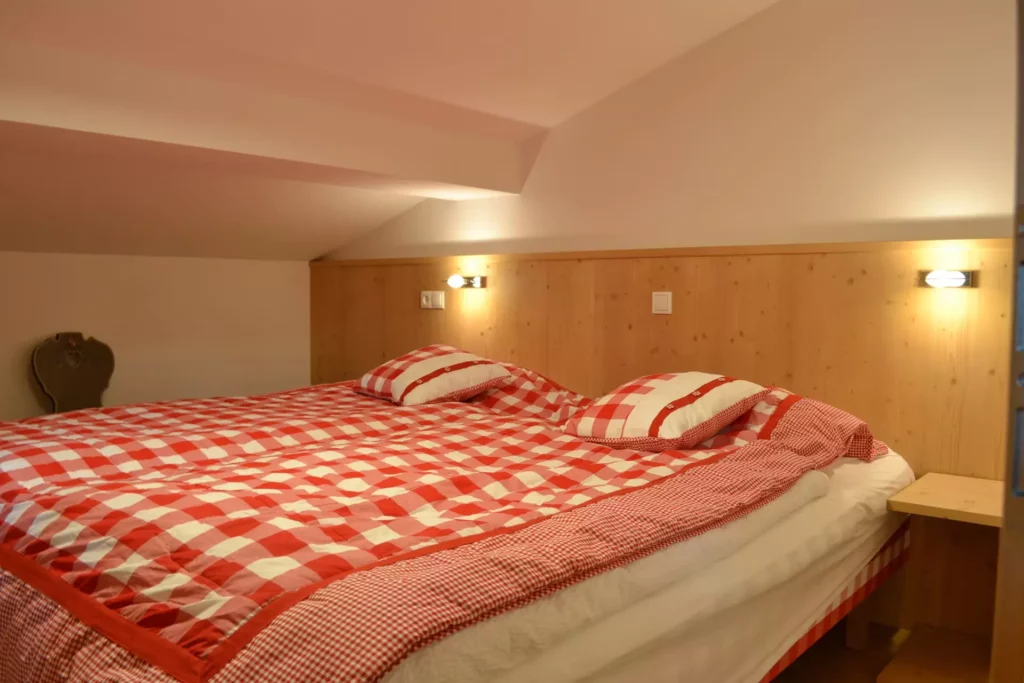 Schlafzimmer mit schräger Decke, Doppelbett mit rot-weiß kariertem Bettzeug, Holzpaneel und Wandlampen, Referenzen