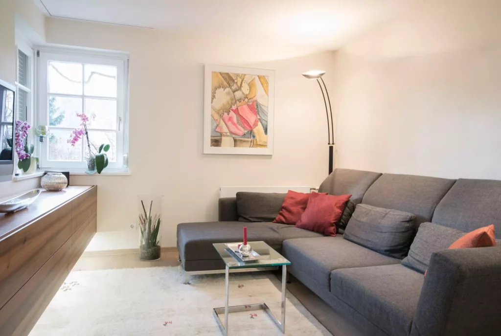 Helles Wohnzimmer mit grauem Poliform Ecksofa, roten Kissen, Glas-Beistelltisch, Holzsideboard, Stehlampe und, Referenzen