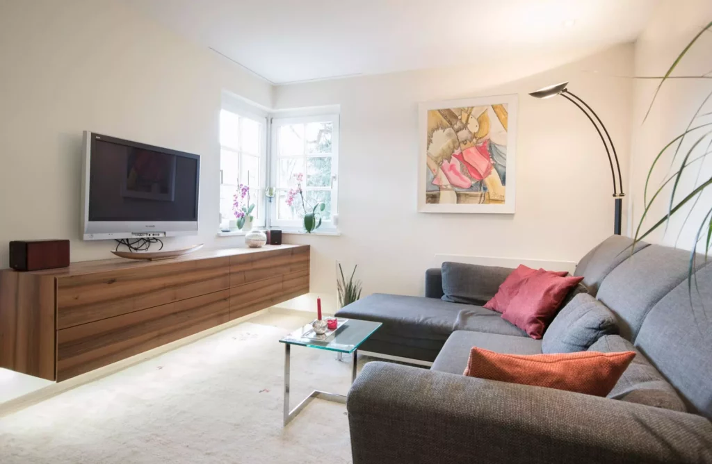 Helles Wohnzimmer mit grauem Polsterecksofa, roten Kissen, Glas-Couchtisch, hängendem Holzsideboard und, Referenzen