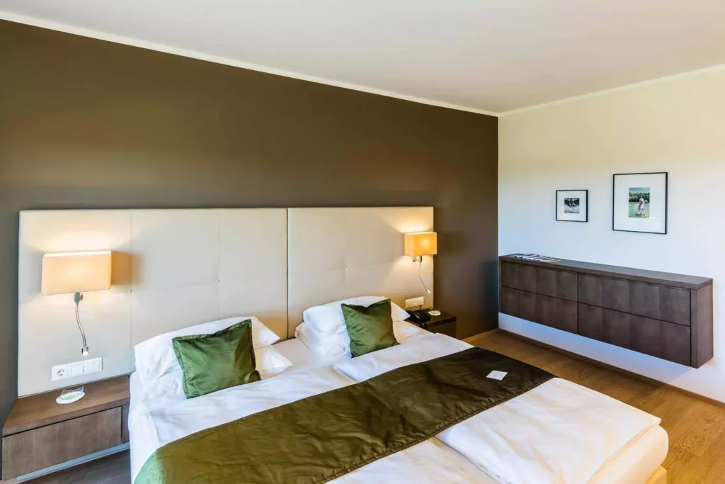 Modernes Hotelzimmer mit Doppelbett, grünem Bettläufer, gepolstertem Kopfteil, Wandlampen und schwebendem, Referenzen