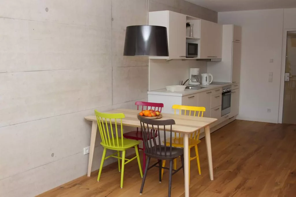 Moderne Küche mit weißer Einbauküche, bunten Stühlen um hellen Holztisch und schwarzer Hängeleuchte vor, Referenzen