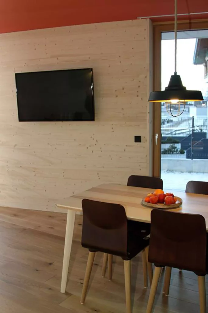 Essbereich mit hellem Holztisch, vier dunklen Stühlen, hängender Lampe, TV an Holzpaneelwand und Fenster mit, Referenzen
