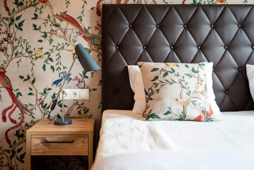 Schlafzimmer mit braunem, gesteppter Kopfteil, Holznachttisch mit grauer Leselampe und Kissen mit floralem, Minotti