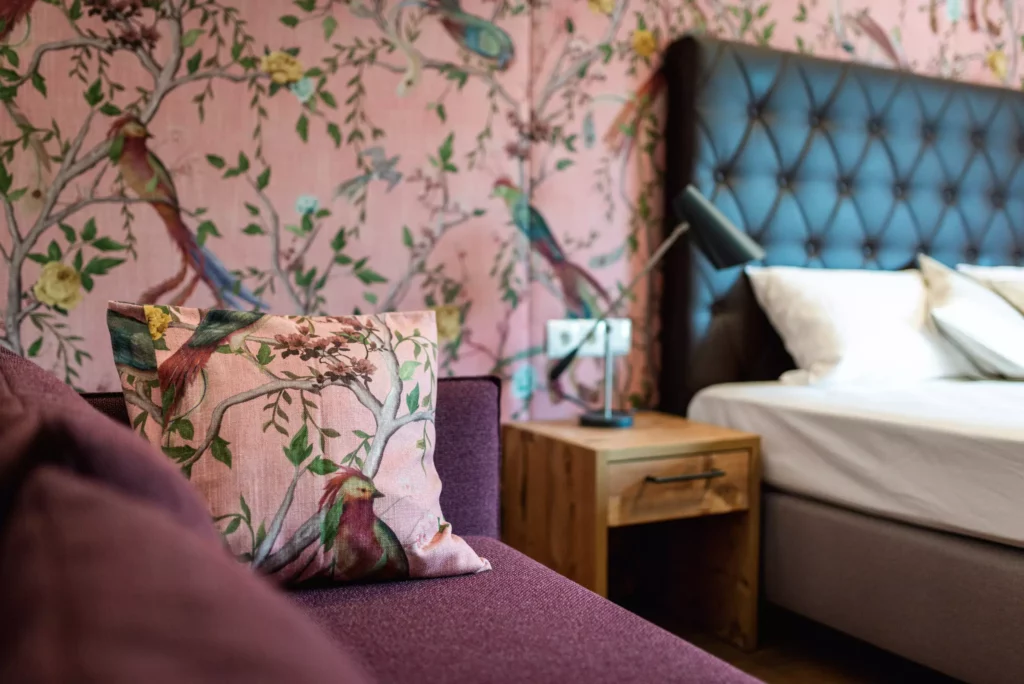 Schlafzimmer mit lila Sofa, Kissen mit Vogelmotiv, Holznachttisch und gepolstertem Minotti-Bett vor floral gemusterter Tapete