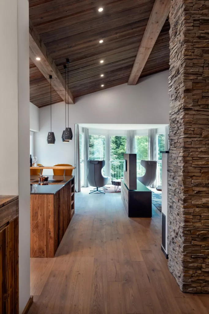 Offener Wohnraum mit Massivholzboden, sichtbaren Holzbalken, moderner Küche, Kamin mit Natursteinverkleidung und Minotti