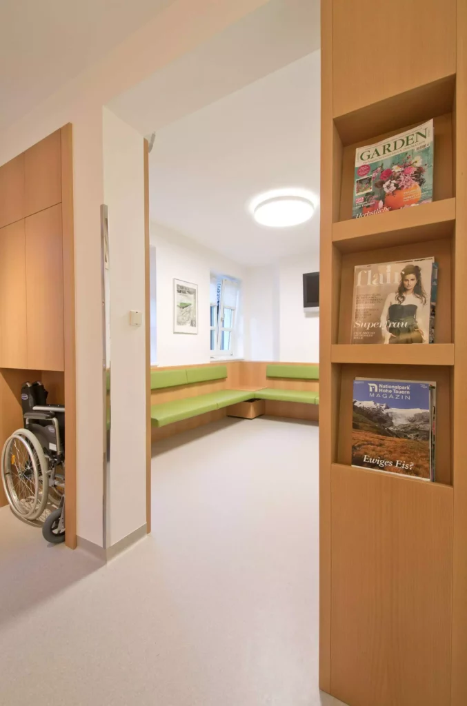 Wartebereich einer Arztpraxis mit maßgefertigten Holzmöbeln, grünen Sitzpolstern, Zeitschriftenregal und Rollstuhl, Referenz