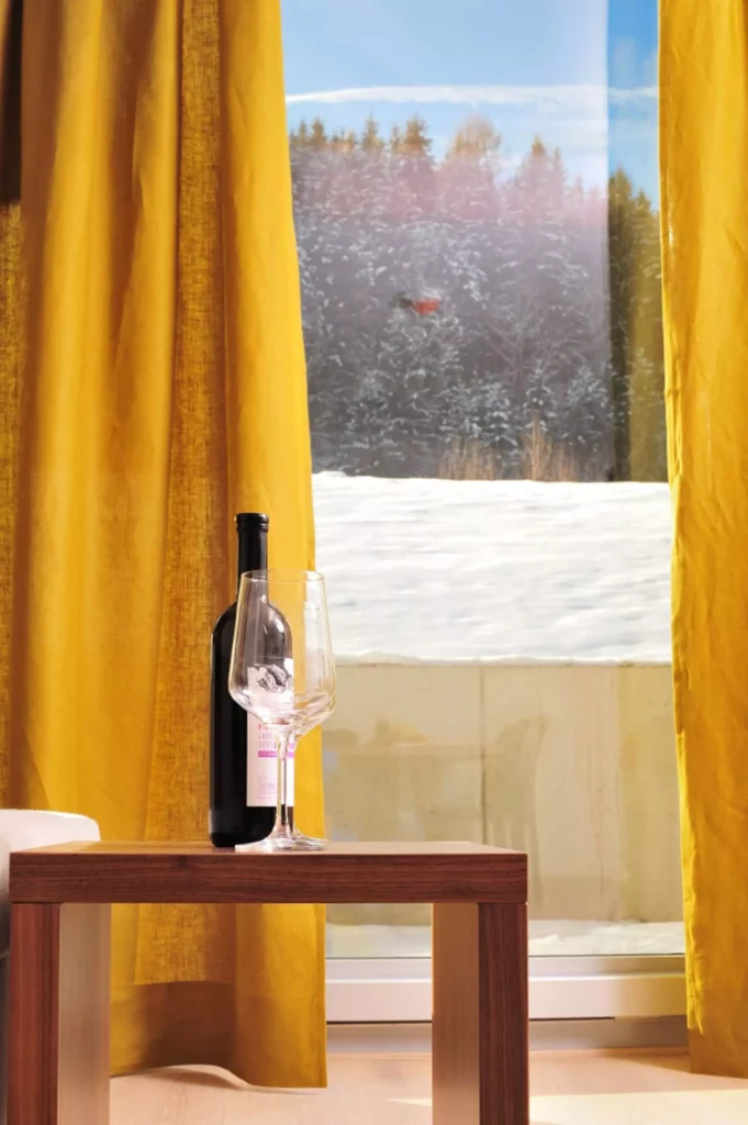 Holztisch mit Weinflasche und Weinglas vor gelben Vorhängen und verschneiter Winterlandschaft im Tauern Residence, Referenz
