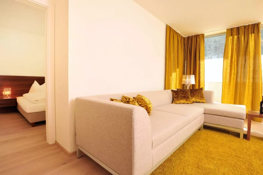 Helles Wohnzimmer mit beigem Ecksofa, goldenen Samtkissen, gelbem Teppich und Vorhängen, Blick in Schlafzimmer mit, Referenz