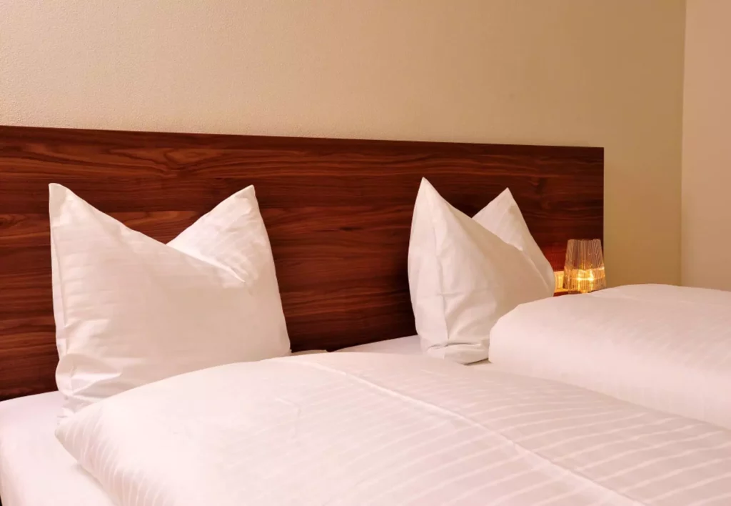 Doppelbett mit weißer Bettwäsche und Kissen vor einem Holz-Kopfteil, dezente Beleuchtung im Tauern Residence, Referenz
