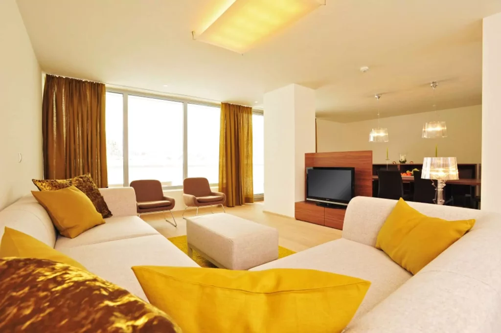 Wohnzimmer mit beiger Polsterecke und gelben Kissen, zwei Sesseln, TV-Möbel aus Holz und Essbereich im, Referenz