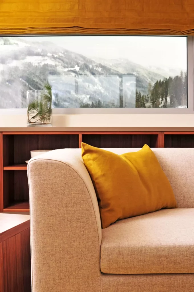 Beiger Polstersessel mit senfgelbem Kissen vor Holzregal und großem Fenster mit Bergblick in der Tauern Residence, Referenz