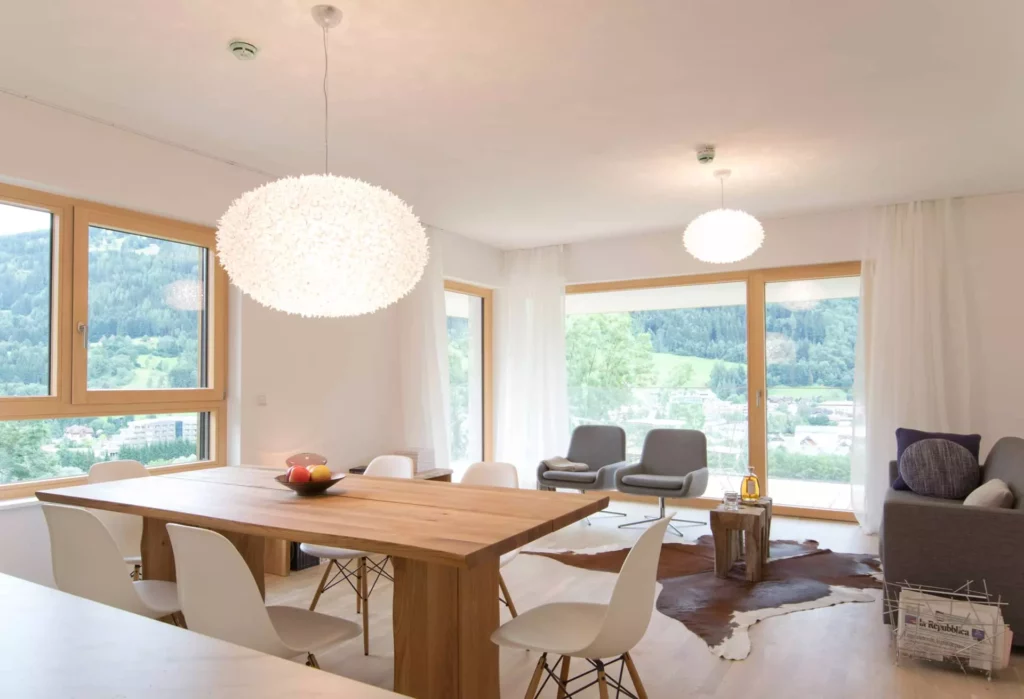 Helles Wohnzimmer mit großem Holztisch, weißen Stühlen, zwei grauen Sesseln, Sofa, Fellteppich und großen Fenstern, Referenz