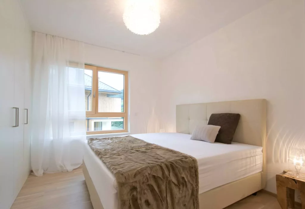 Helles Schlafzimmer mit Polsterbett, zwei Kissen, braunem Fellüberwurf, Gardine, Fenster und Holzboden in, Referenz