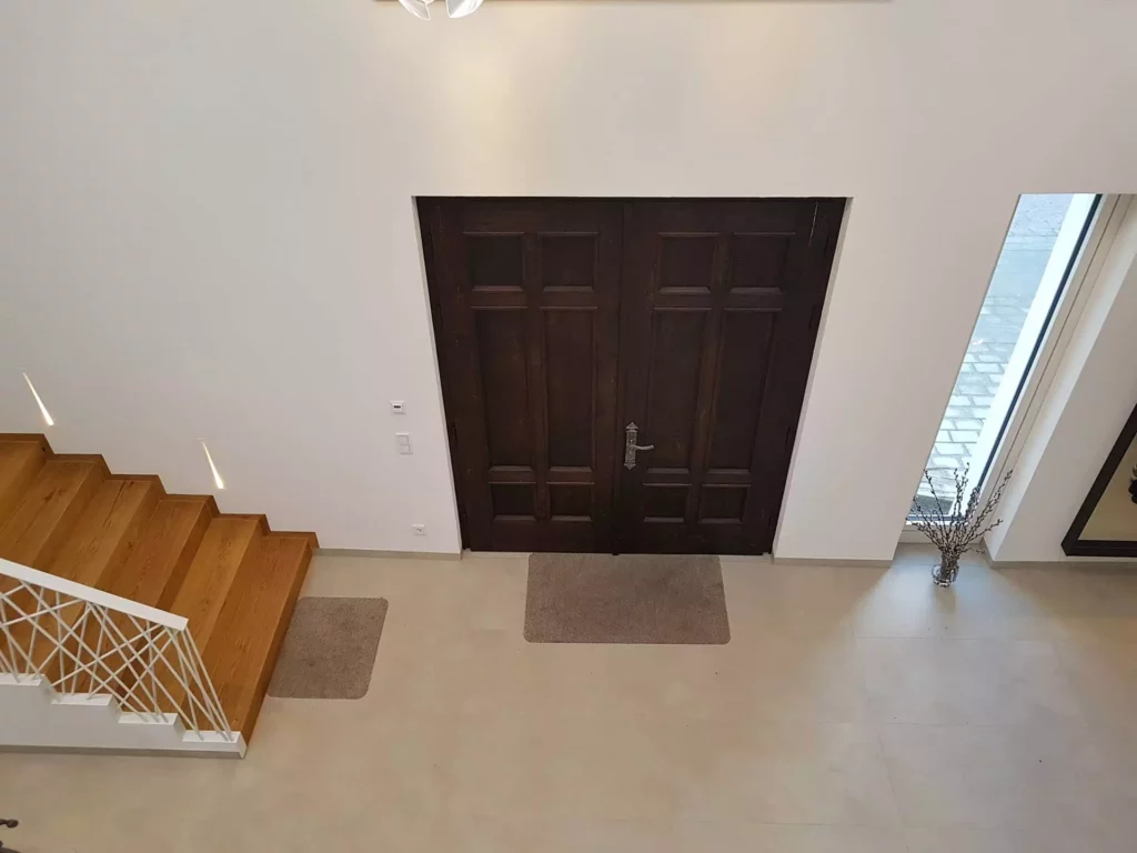 Eingangsbereich einer Villa mit dunkler Holztür, heller Wand, moderner Holztreppe mit weißem Geländer und großem, Referenz