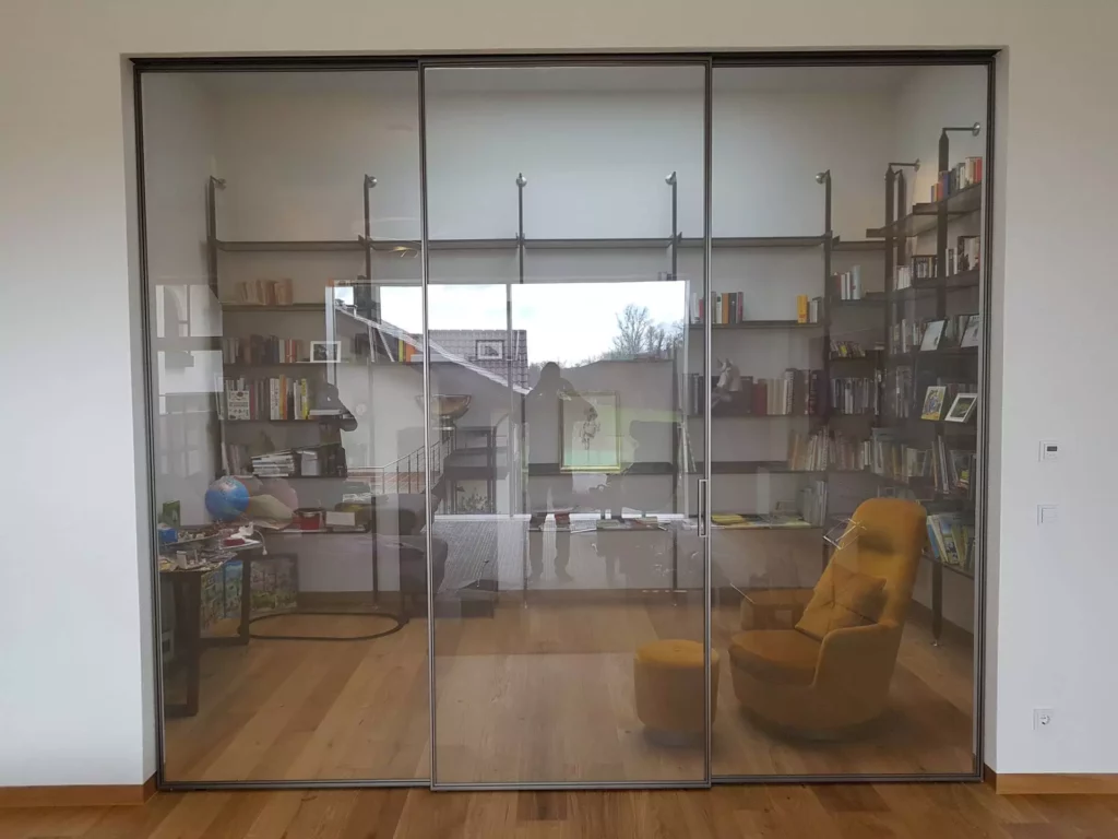 Glas-Schiebetür mit Rahmen als Raumtrennung zu einem Leseraum mit Wandregal, gelbem Sessel und Hocker, Holzfußboden, Referenz
