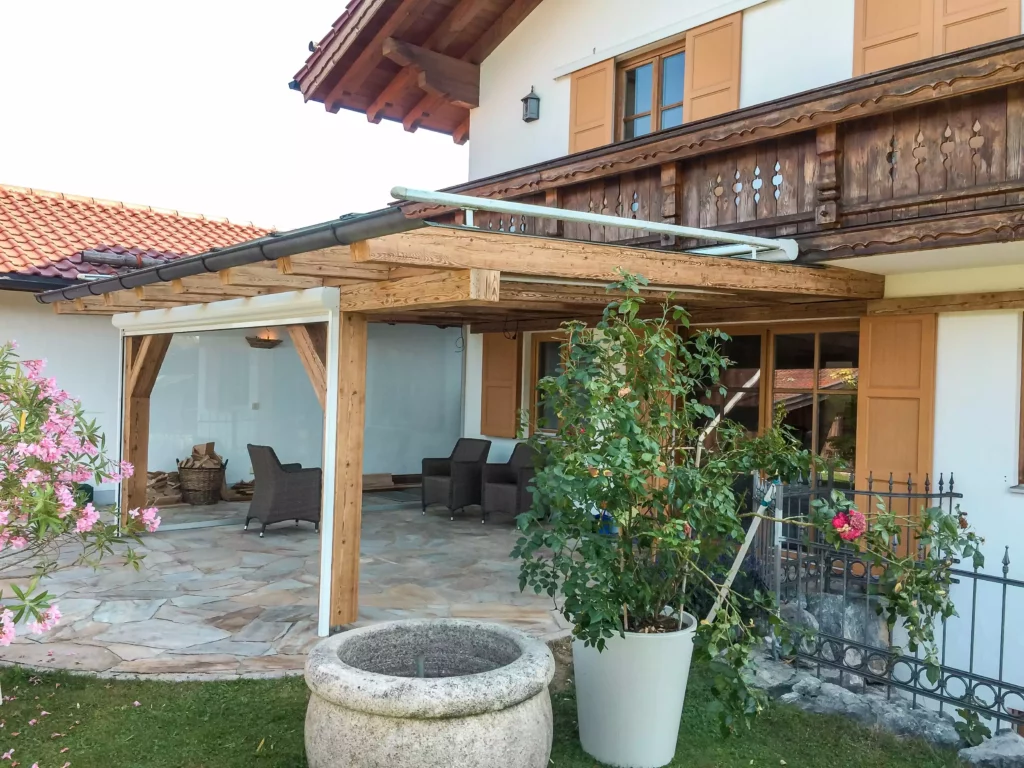 Terrasse mit Altholzüberdachung und Sitzbereich vor einem Haus mit Holzfenstern und Balkon in traditioneller, Altholzmöbel