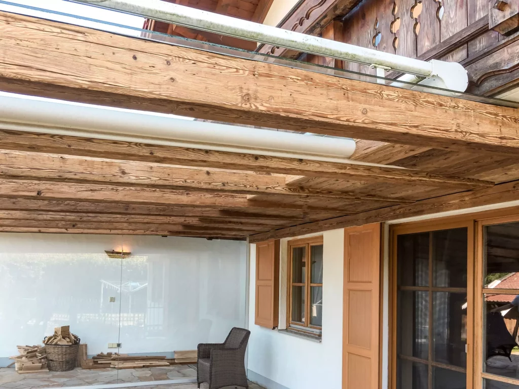Überdachte Terrasse mit Altholzdecke, Glaswand, Holzfenstern mit Klappläden, Korbsessel und Holzstapel im, Altholzmöbel